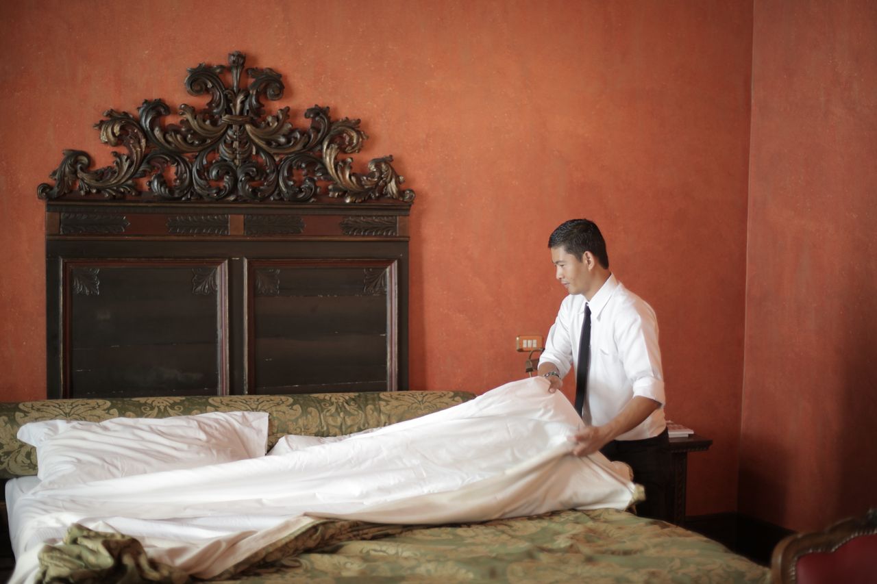 heros-img A hotel staff member in formal attire making a bed in a luxurious bedroom setting.