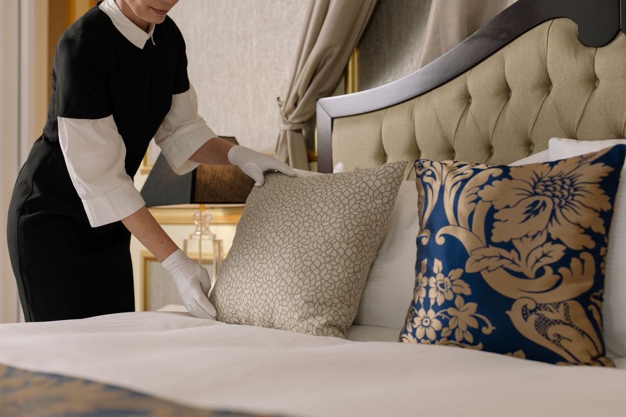about-us A chambermaid arranges pillows in a luxurious hotel room, showcasing comfort and elegance.